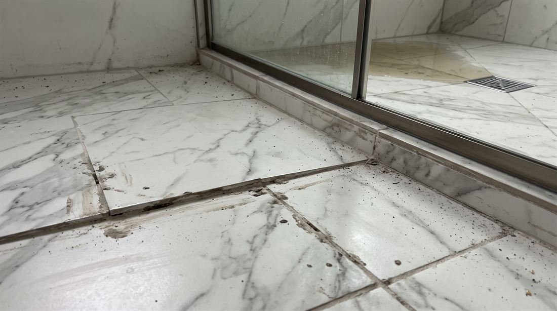 Piso de porcelanato recém-assentado, torto e com desnível visível, rejunte manchado e poça de água parada longe do ralo